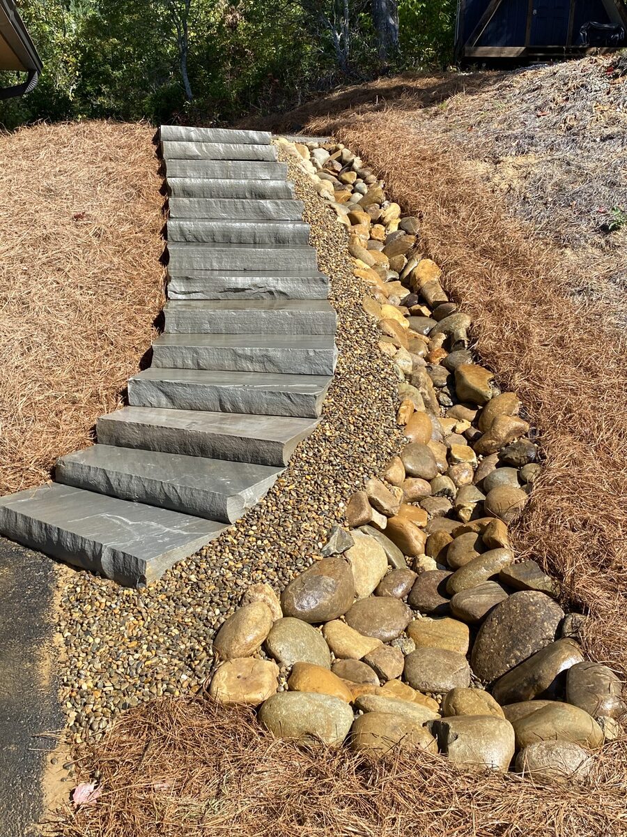 Natural stone slab steps beside a river-rock drainage channel on a pine-strawed hillside